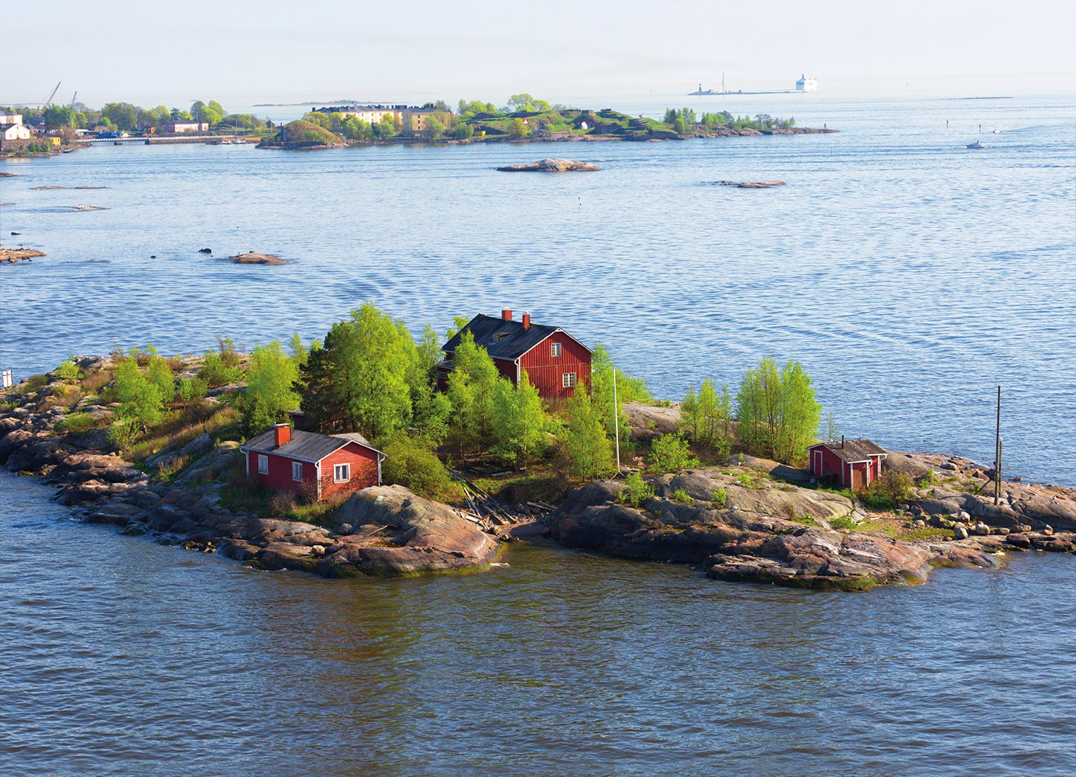 Kleine licht-bewaldete Felseninsel mit zwei roten Holzhäusern  im Hintergrund Hafen und Industrie